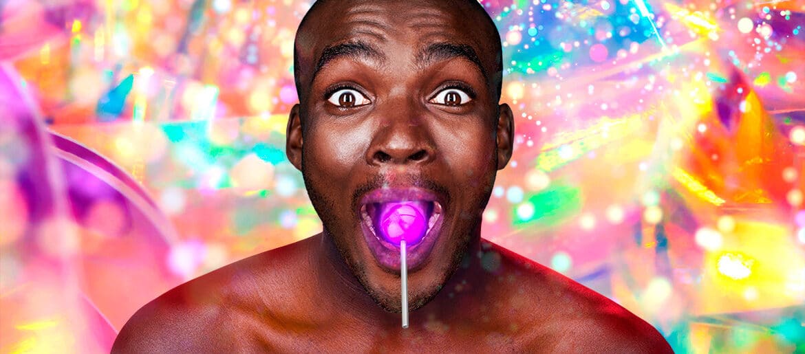 Close up of a smiling, wide-eyed, bare-chested black man, with a glowing purple lollipop in his mouth. The background behind him is bright, multi-coloured and full of sparkles