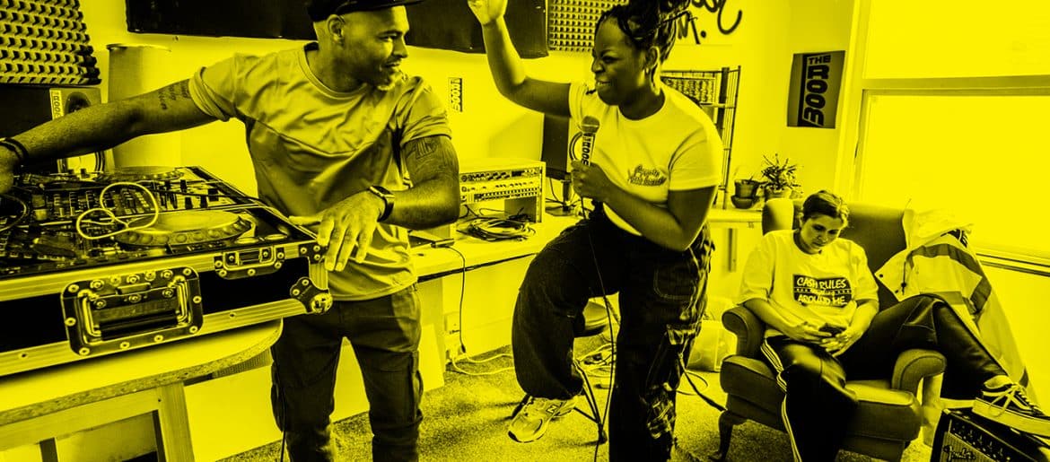 Three people in a room DJing, holding a microphone and sitting looking at their mobile phone